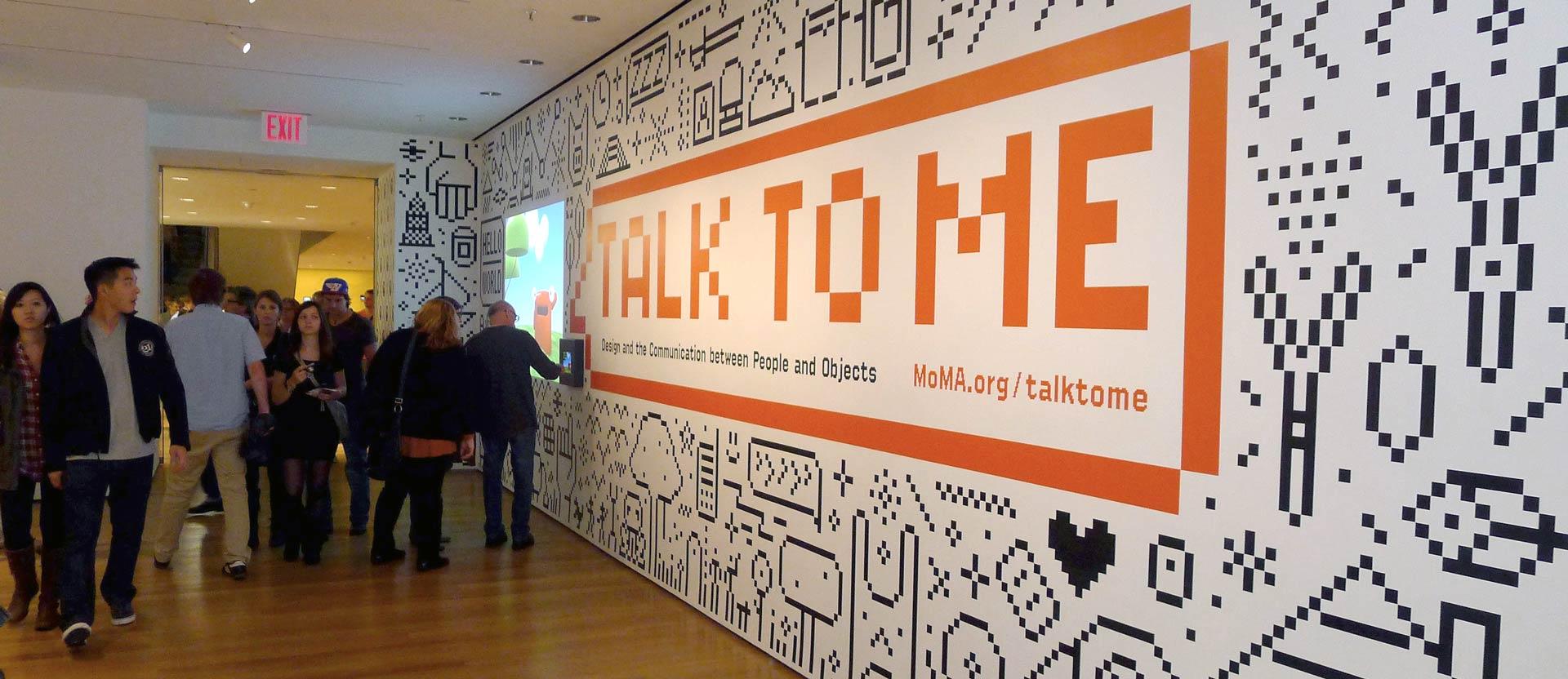 The entrance wall of the Talk to Me exhibition at the Museum of Modern Art: an orange TALK TO ME wordmark set into a wall of black-and-white pixel illustrations, with visitors walking past