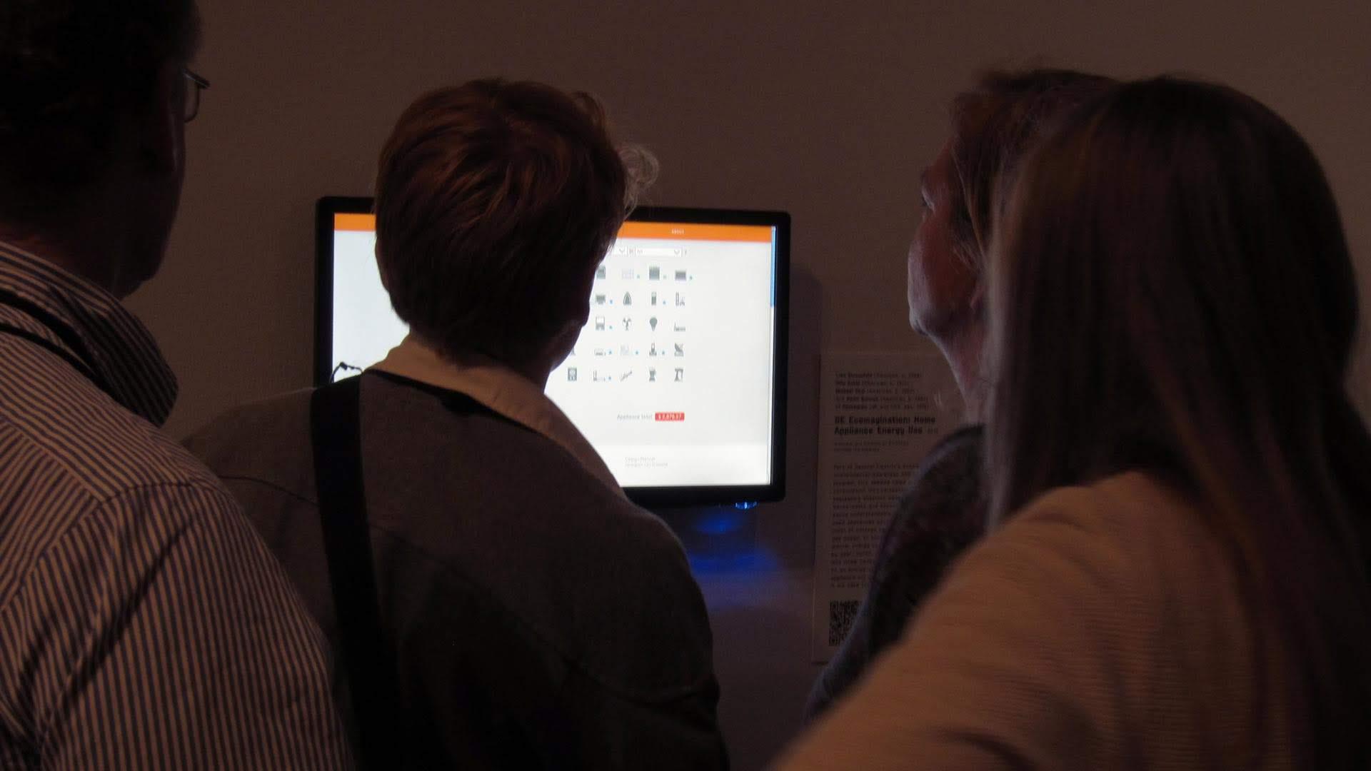 Visitors at the Museum of Modern Art standing at a monitor running the Home Energy Calculator, the wall label beside it